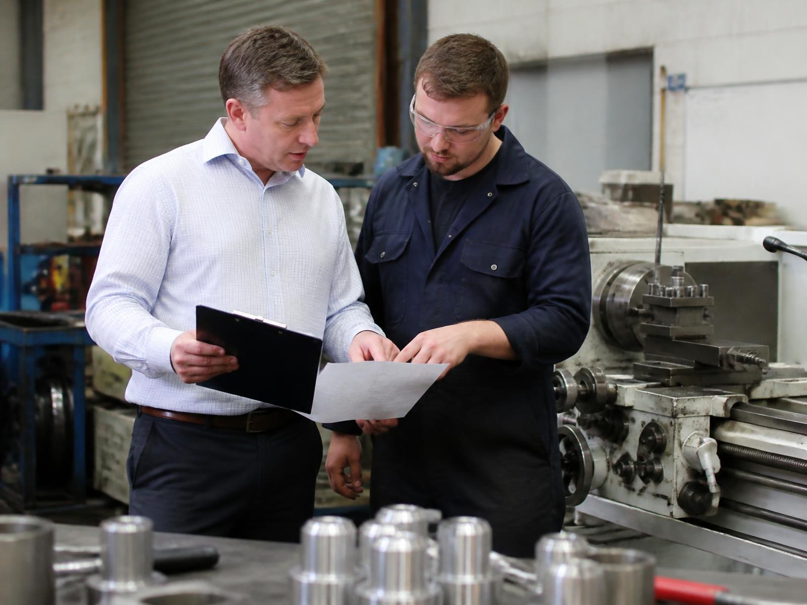 An ISO consultant reviewing technical drawings with an engineer beside a lathe in a precision engineering workshop